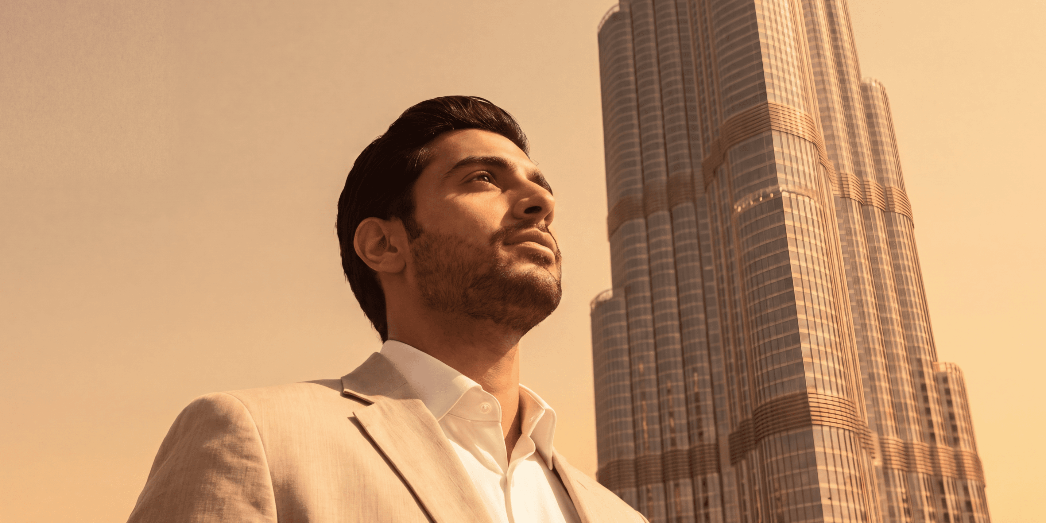 Man in a light beige suit and white shirt looking upward, standing before the Burj Khalifa at golden hour, Dubai.
