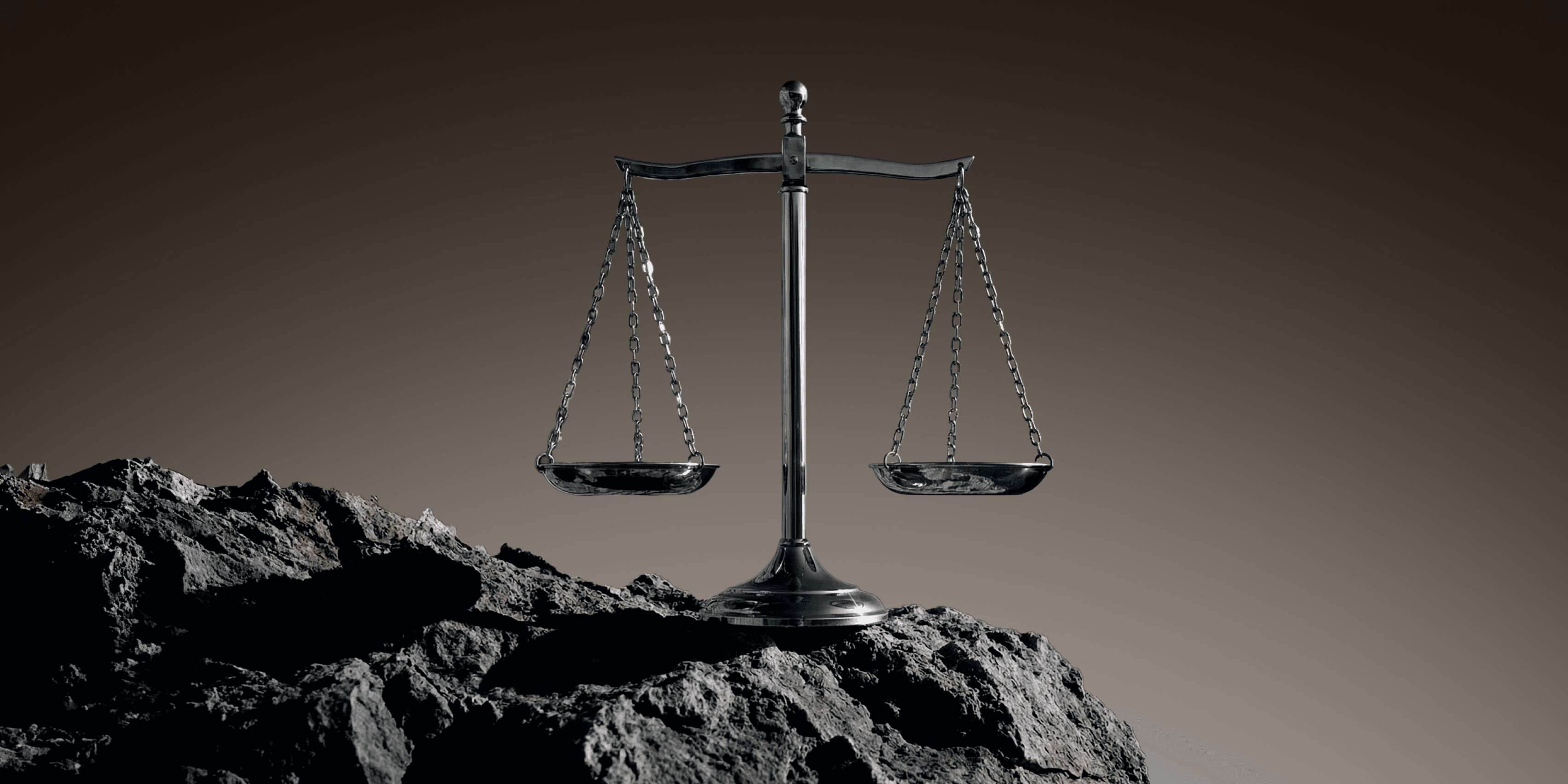 A metal balance scale with two empty pans, standing on dark rocky ground against a brown background.