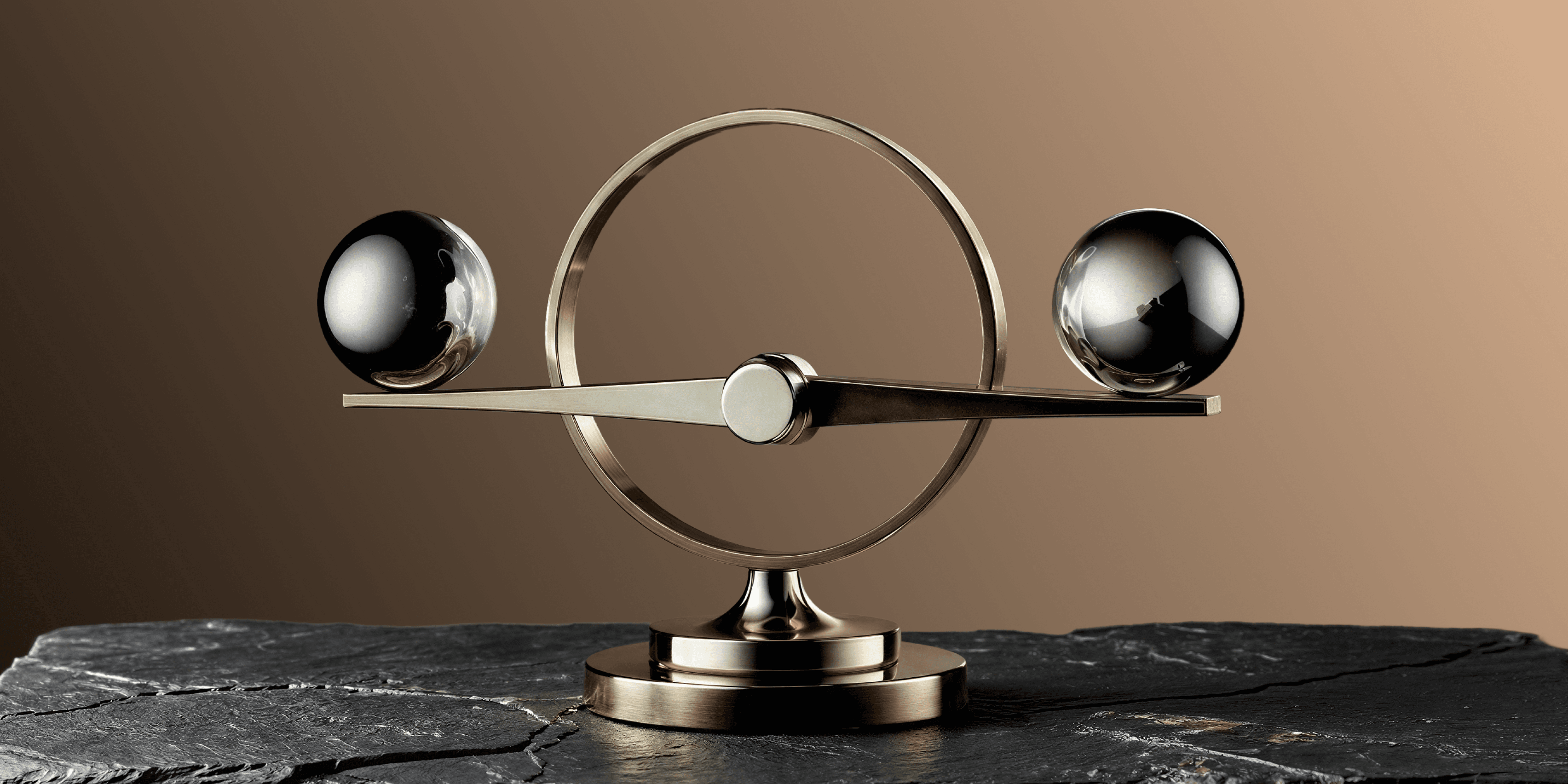 A brass and chrome balance sculpture with two polished metal spheres suspended on a horizontal beam within a circular frame, mounted on a pedestal atop dark slate stone, set against a warm brown gradient background.