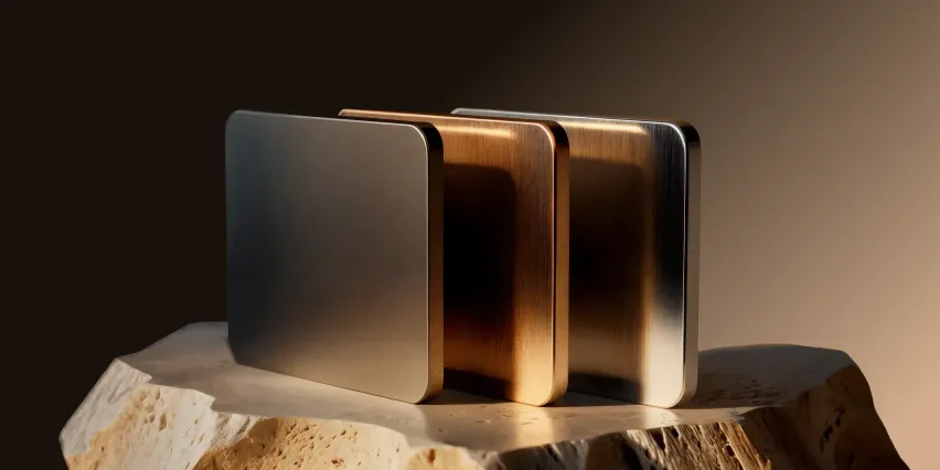 Three upright metal plates in brushed silver, bronze and steel standing side by side on a rough pale stone, lit warmly against a dark brown background