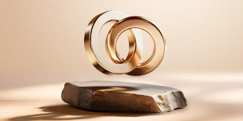 Two interlocking gold rings, one pale and one warmer rose gold, floating above a rough stone slab against a soft beige background