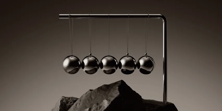 Newton's cradle with five polished chrome balls hanging from a metal frame, resting on a jagged rock against a dark brown background