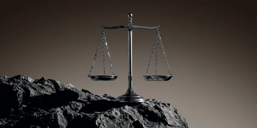 A metal balance scale with two empty pans, standing on dark rocky ground against a brown background.