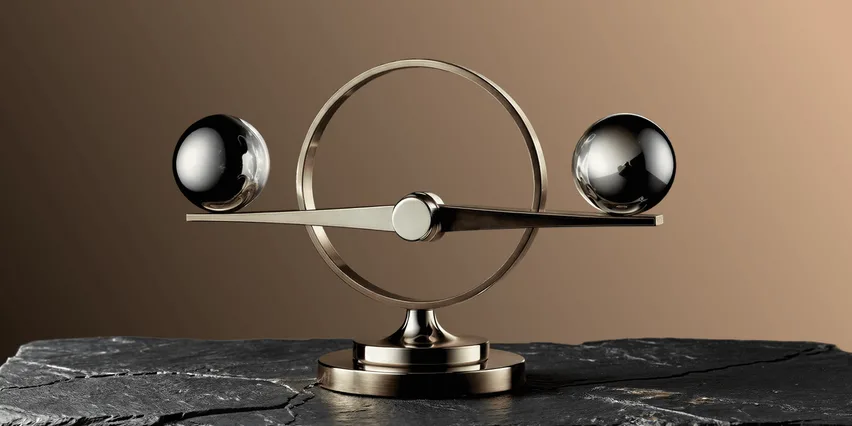 A brass and chrome balance sculpture with two polished metal spheres suspended on a horizontal beam within a circular frame, mounted on a pedestal atop dark slate stone, set against a warm brown gradient background.