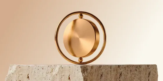 Brushed gold disc suspended inside a thin gold ring, balanced on a rough travertine stone block against a warm beige background.