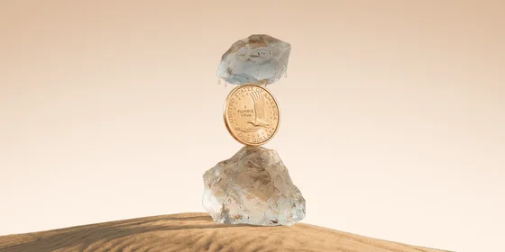 A gold US one-dollar coin standing on edge, wedged between two blocks of melting ice on a sand dune, with meltwater dripping down onto it against a warm beige background.