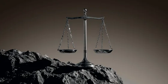A metal balance scale with two empty pans, standing on dark rocky ground against a brown background.