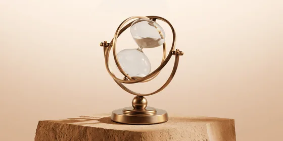 Brass hourglass suspended in gimbal rings on a sandstone block, sand running through against a warm beige background