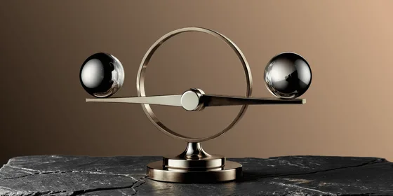 A brass and chrome balance sculpture with two polished metal spheres suspended on a horizontal beam within a circular frame, mounted on a pedestal atop dark slate stone, set against a warm brown gradient background.